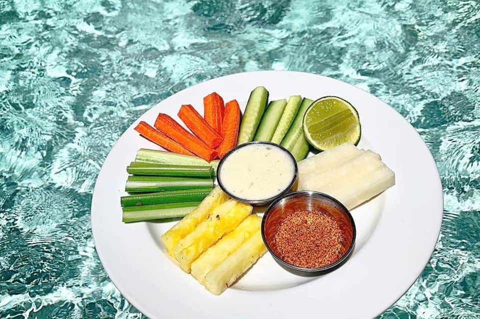 White platter of fresh vegetable crudités with pineapple, carrots, cucumbers, and dipping sauces poolside