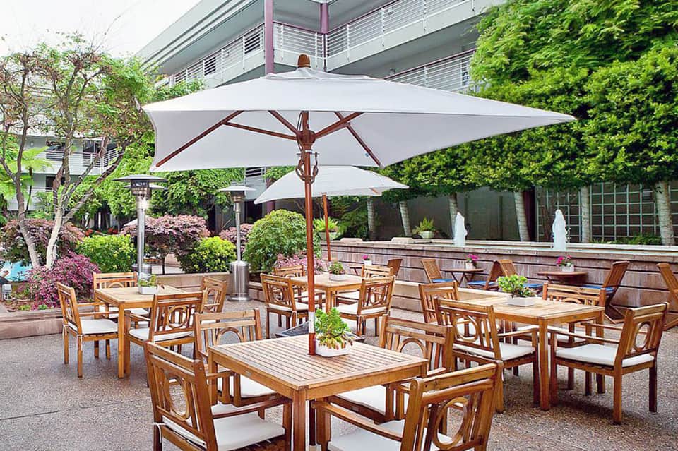 Outdoor patio dining area with wooden tables and chairs under large white umbrellas and green landscaping