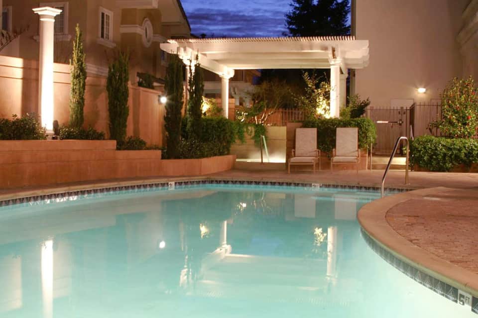 Illuminated resort pool at dusk with white pergola, lounge chairs, and manicured landscaping
