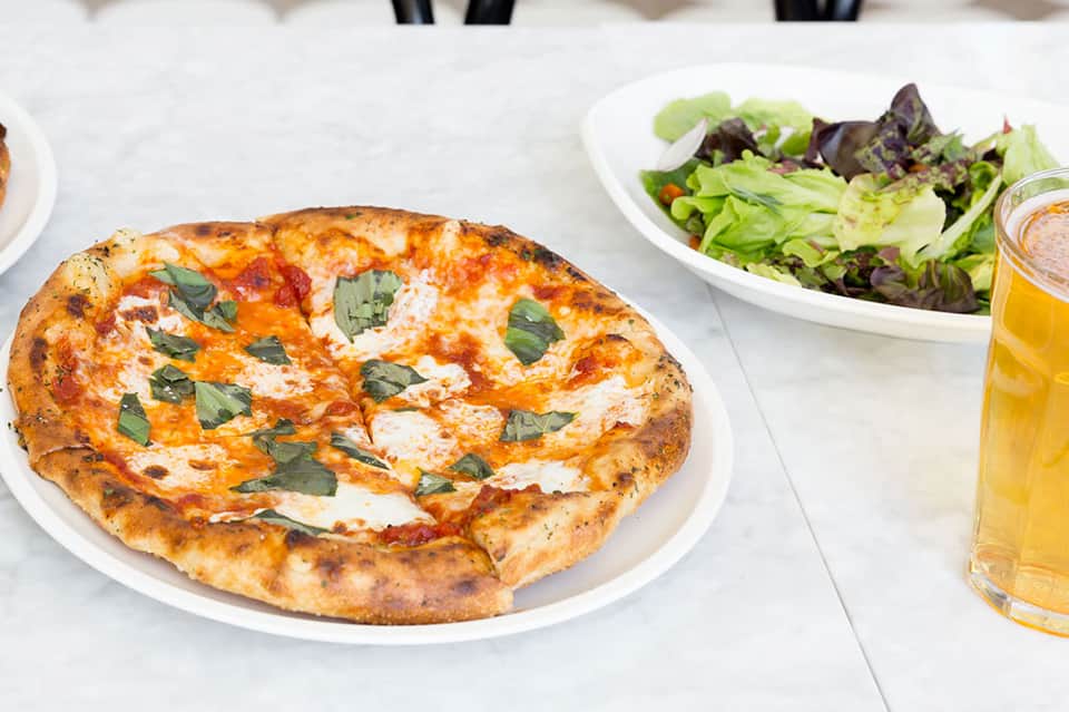 Margherita pizza with fresh basil on white plate, green salad and beverage in background