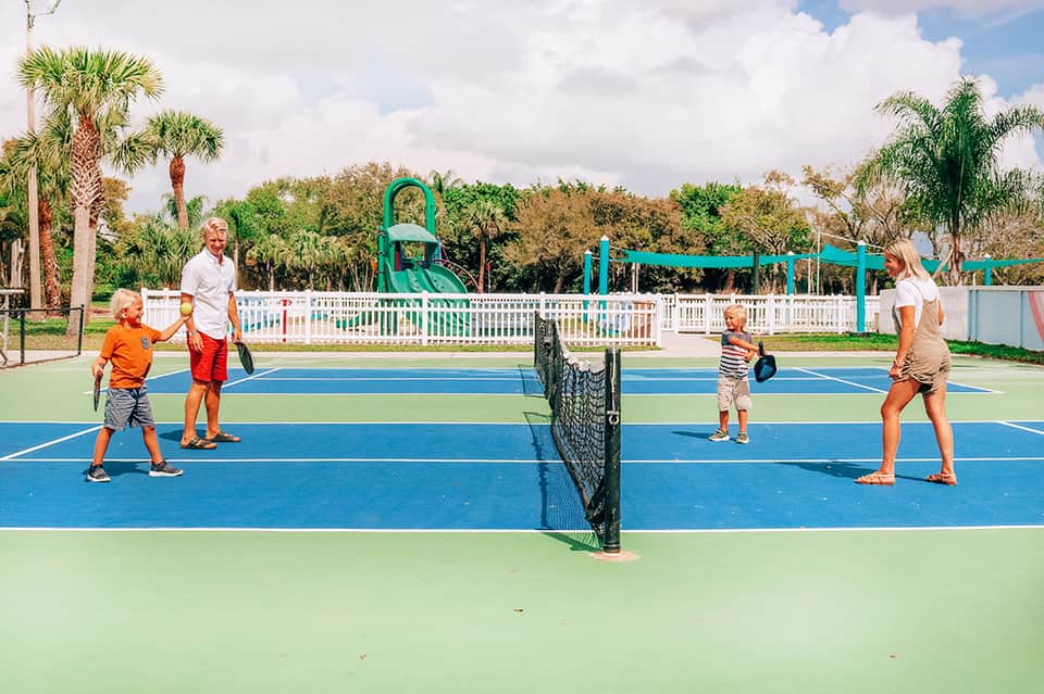Family playing pickleball on an outdoor court with white railings and palm trees