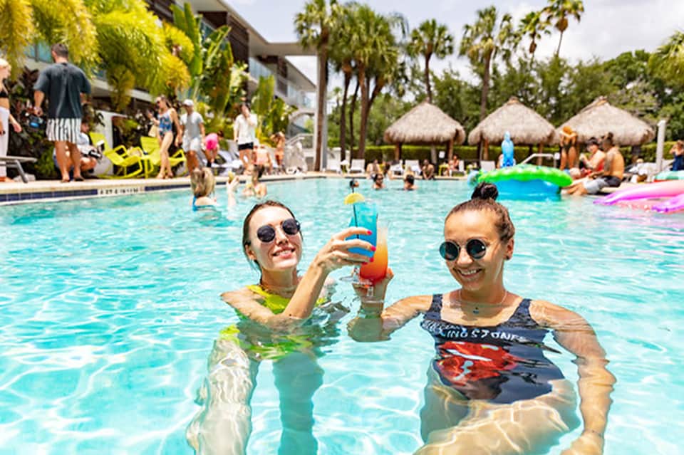 Two guests swimming in bright blue pool with floats, palm trees and thatched umbrellas visible