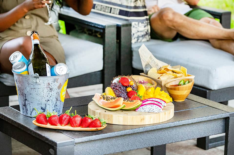 Outdoor dining setup with fruit platter, snacks, ice bucket with beverages, and guests relaxing nearby
