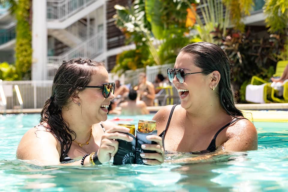 Two women in pool holding drinks, smiling, with resort buildings and sunbeds in background