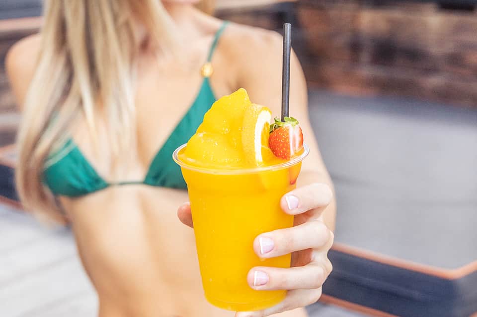 Woman in teal bikini holding tropical yellow drink with fruit garnish at beach