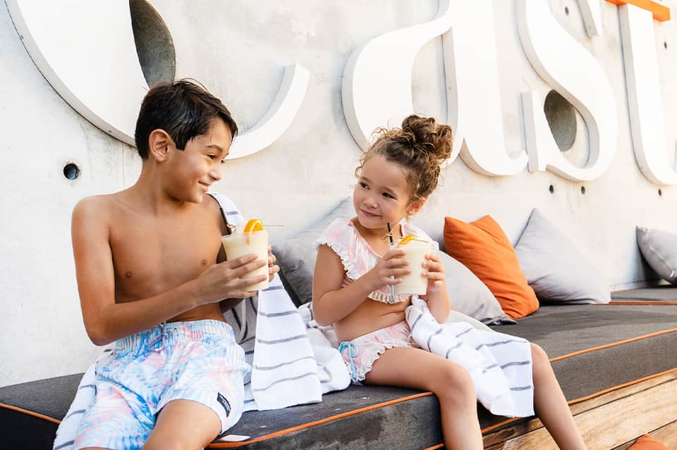 Two children enjoying beverages poolside against white wall with decorative circular design