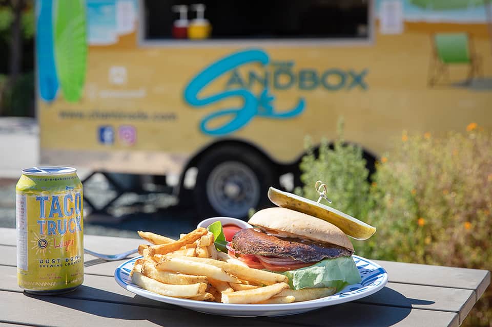 Gourmet burger with fries and beverage in front of Sandbox food truck branding