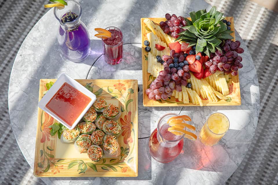 Overhead view of poolside table with fruit platters, appetizers, and tropical beverages in sunlight