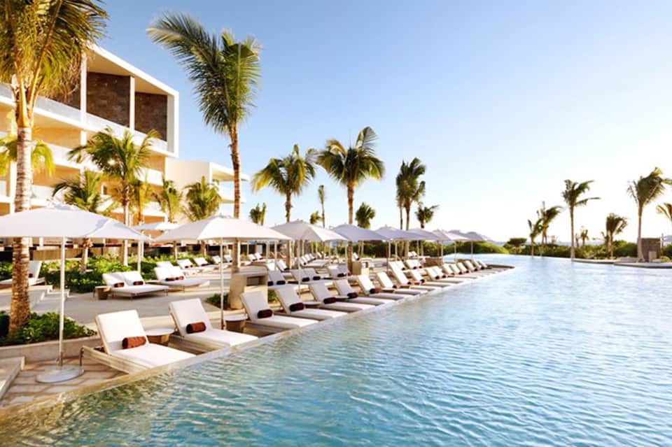 Resort infinity pool with white loungers, palm trees, umbrellas, and mountains in background