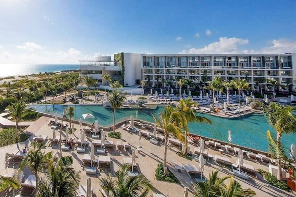Beachfront resort with modern white building, turquoise lagoon pool, palm trees, and ocean view under clear sky.