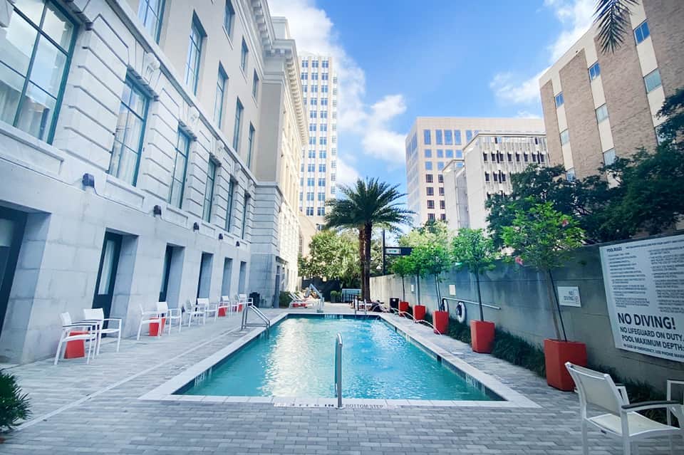 Urban rooftop pool surrounded by tall buildings with lounge chairs and red umbrellas