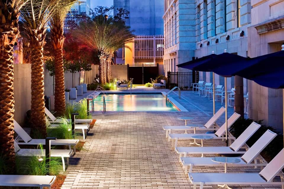 Illuminated resort pool deck at dusk with palm trees, lounge chairs, and blue umbrellas