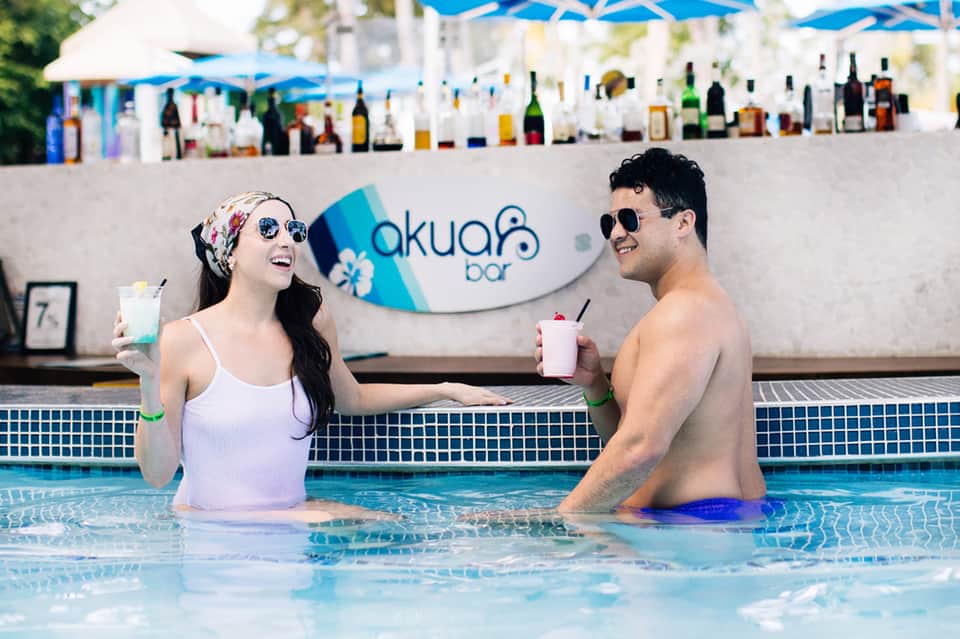 Couple enjoying cocktails in pool at Akua Bar with blue umbrellas and bar visible