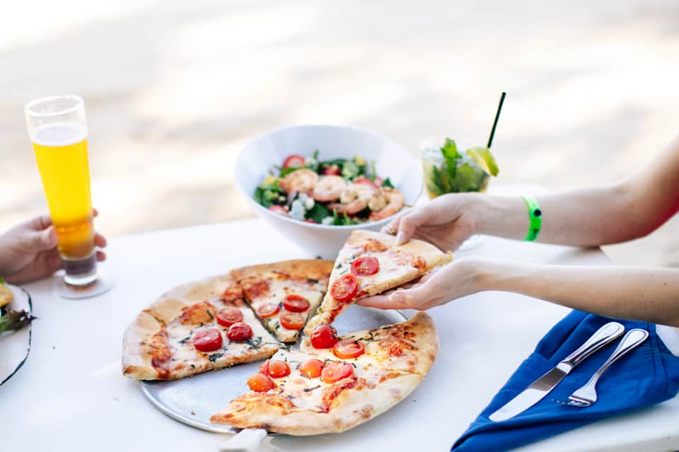 Beach lunch scene with guests enjoying pizza, salad, and drinks at white outdoor dining table