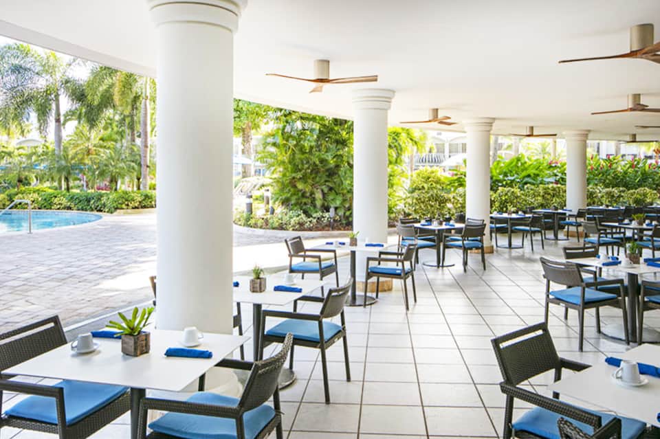 Modern covered dining pavilion with white columns, blue cushioned chairs, and pool view through palm trees