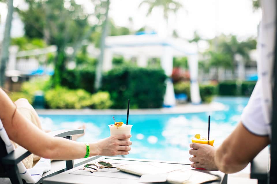 Two people holding tropical beverages overlooking resort pool and gardens
