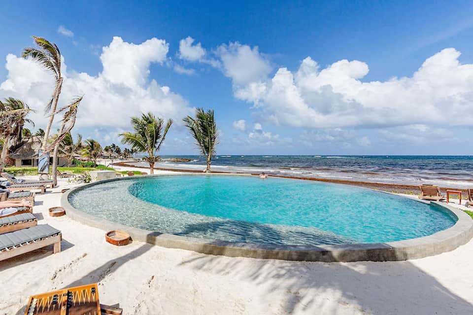 Circular beachfront pool with turquoise water, palm trees, ocean view, and lounging areas