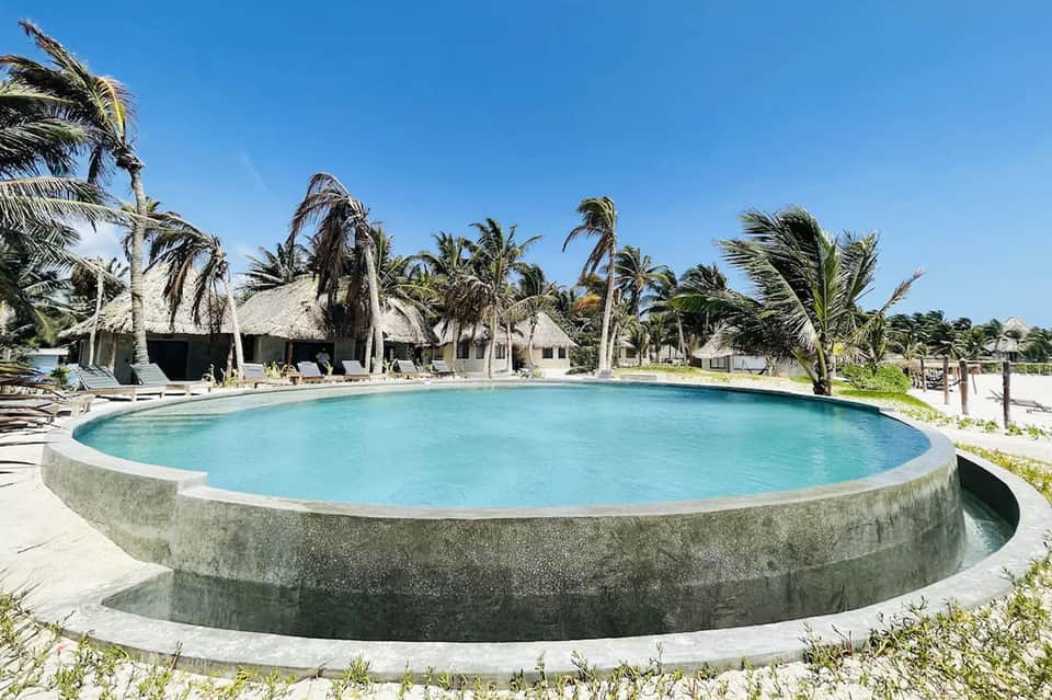 Circular lagoon-style pool surrounded by palm trees with white beach bungalows in background