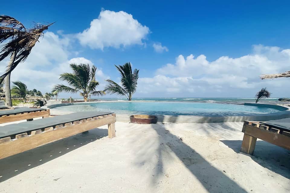 Circular pool with turquoise water surrounded by palm trees and white lounge chairs on white sand