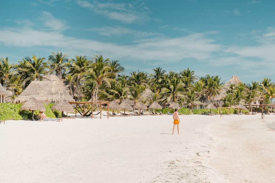 Beach scene with thatched-roof structures, palm trees, white sand, and person in yellow shorts