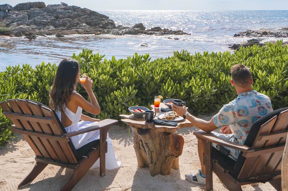 Couple dining at beachfront table with ocean, rocky shoreline, and tropical mangrove vegetation in background