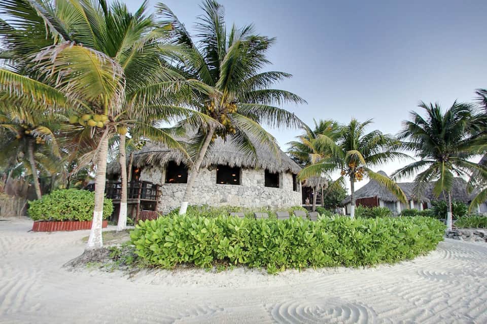 Traditional thatched-roof stone buildings on sandy beach with palm trees