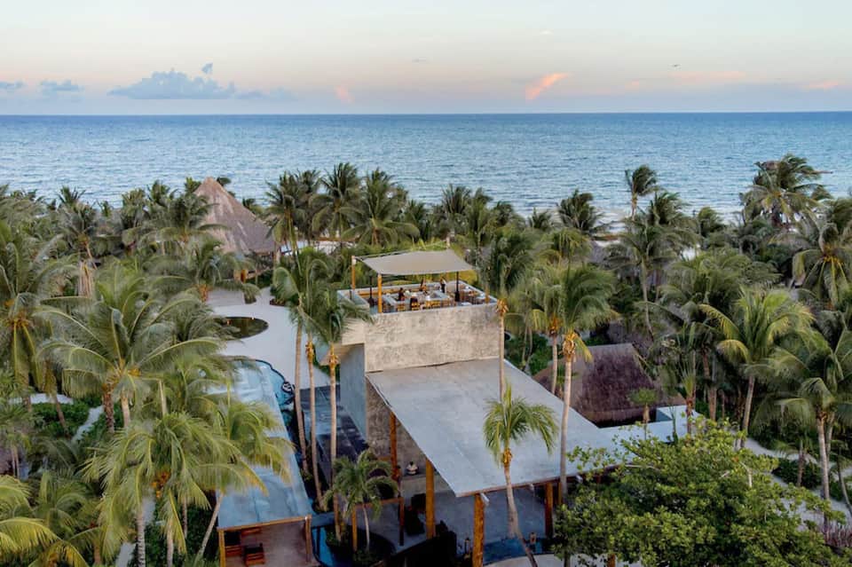 Aerial view of beachfront resort with multi-level structures, pools, and palm trees by ocean