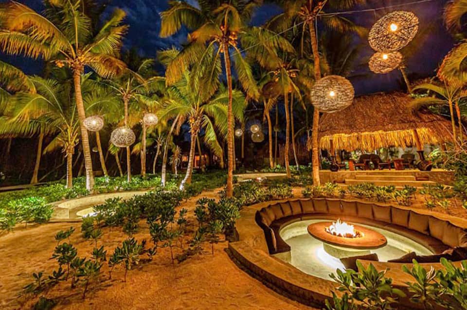 Nighttime beach bonfire area with palm trees, illuminated decorative spheres, thatched-roof structures, and seating