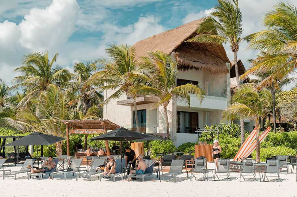 Beachfront resort with modern architecture, thatched roof cabanas, palm trees, and guests lounging