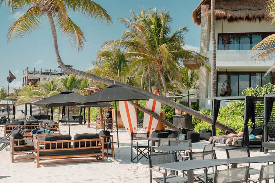 Beach cabana lounge area with wooden furniture, black umbrellas, and palm trees