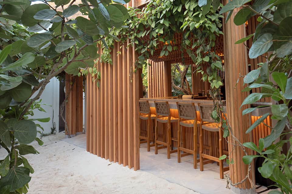 Wooden pergola bar with climbing vines, bar seating, and sand flooring in tropical setting