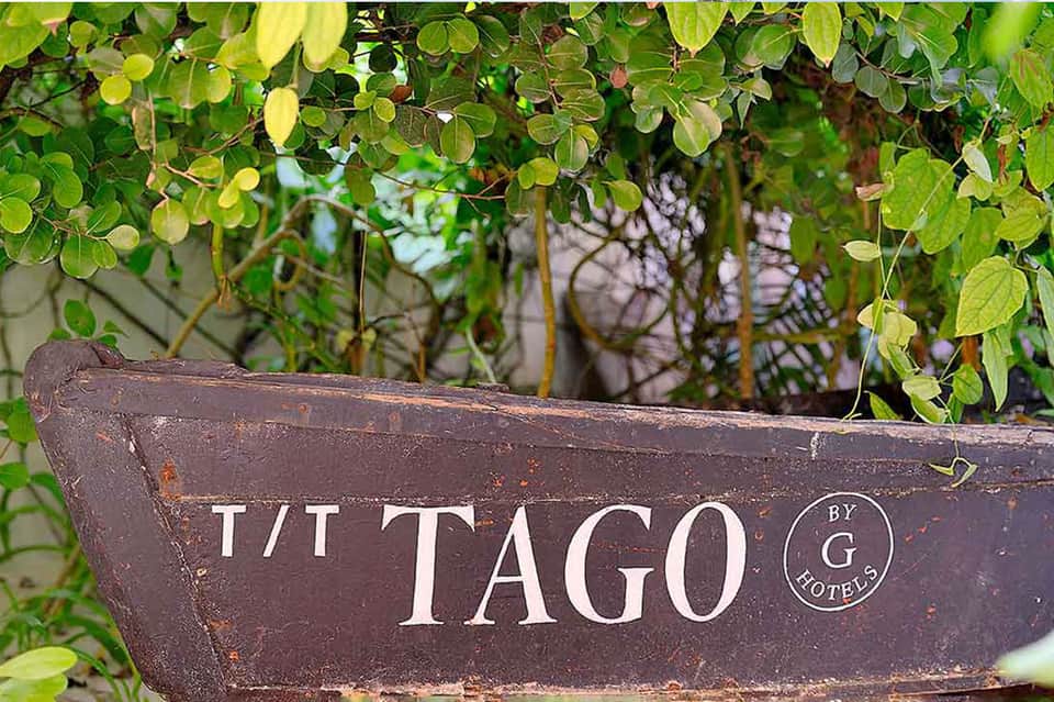 Weathered wooden boat sign for Tago by G Hotels with tropical foliage overhead