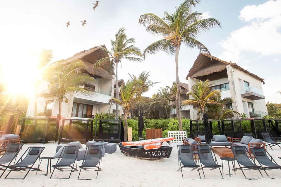Beachfront resort with white buildings featuring thatched roofs, palm trees, and lounge chairs on the sand at sunset.