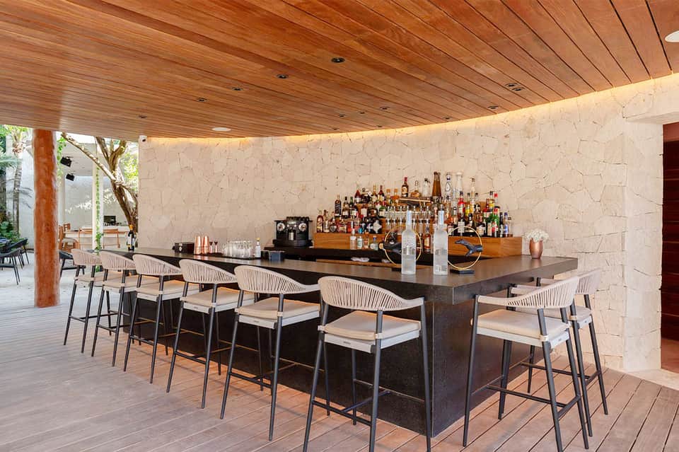 Modern outdoor bar with curved dark counter, rope-detail bar stools, wooden ceiling, and full liquor display