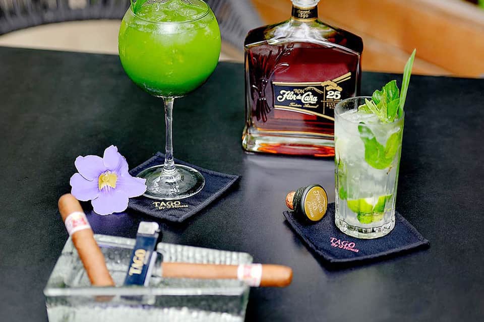 Upscale bar with green and white cocktails, rum bottle, cigars, and decorative purple orchid