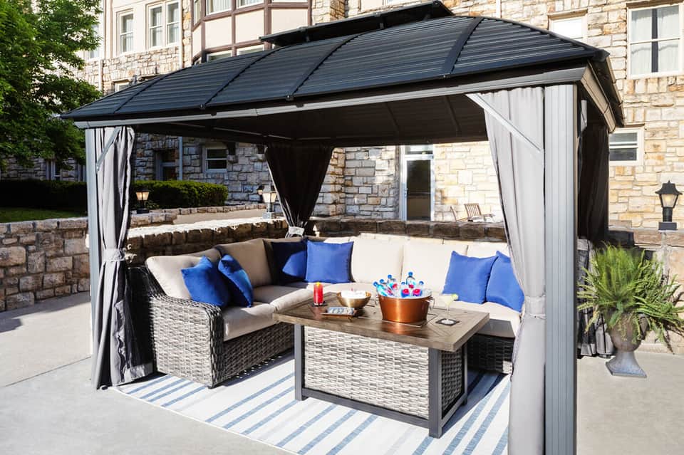Outdoor patio with black metal pergola covering wicker seating, blue pillows, wooden table, and stone building backdrop
