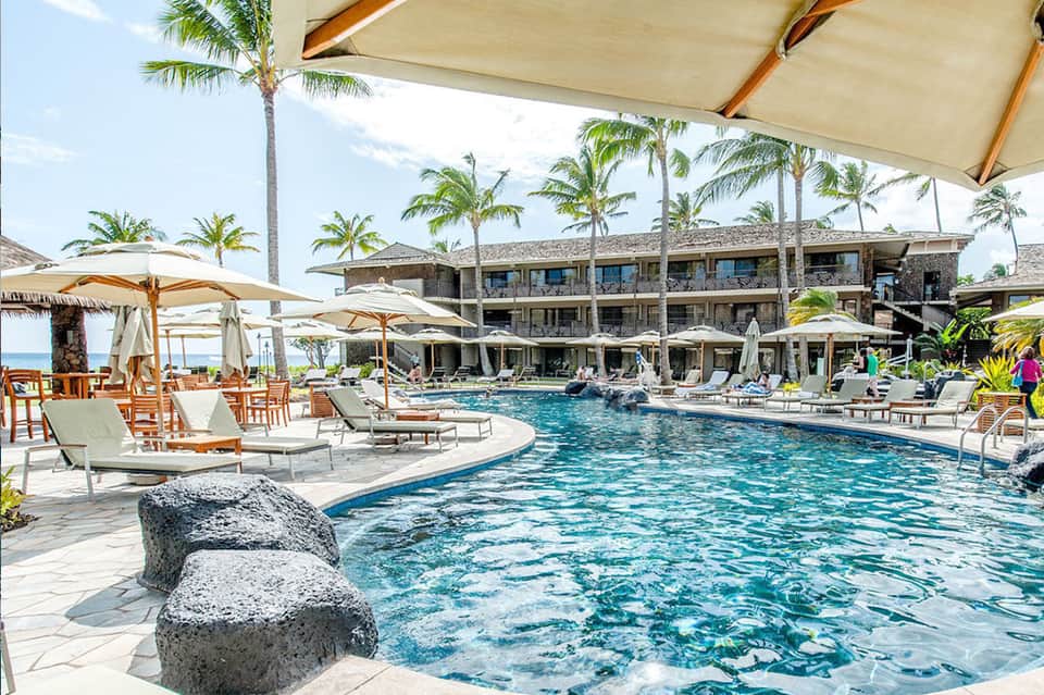 Beachfront resort pool with rock features, loungers, umbrellas, and multi-story stone building