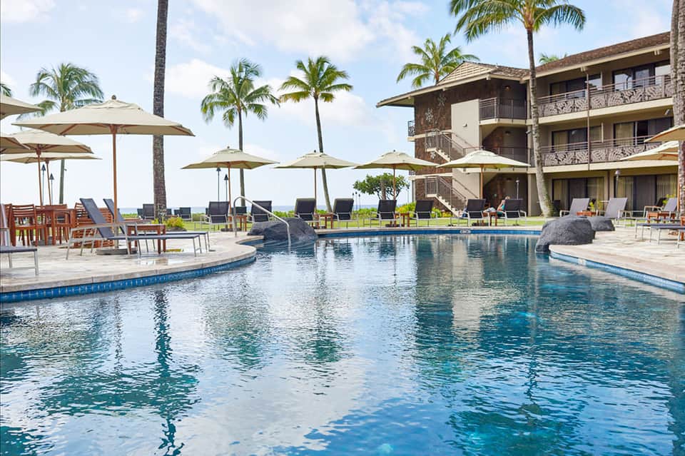 Resort pool with tan umbrellas, lounge seating, palm trees, and ocean view beyond