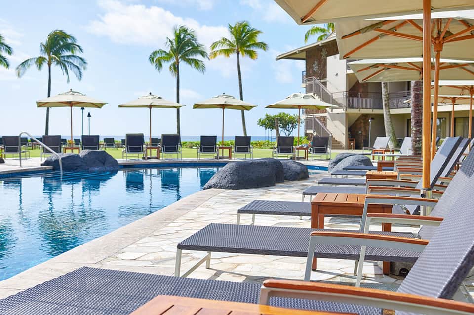 Beachfront resort pool with large rock features, umbrellas, and ocean view with palm trees