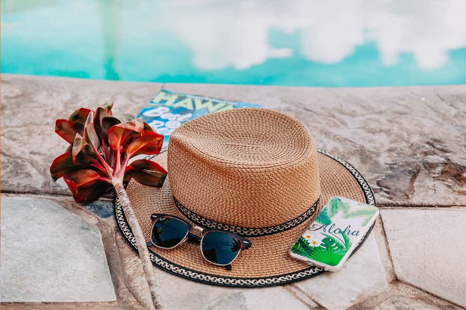 Straw hat, sunglasses, and red flower on pool deck with turquoise water