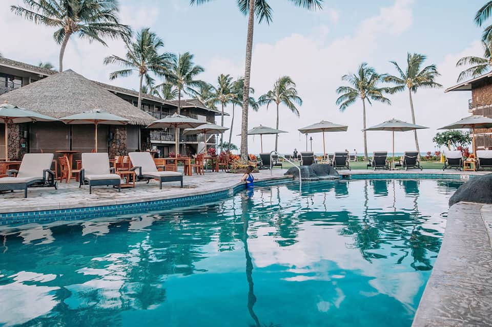 Resort pool area with lounge chairs, thatched cabanas, palm trees, and ocean view