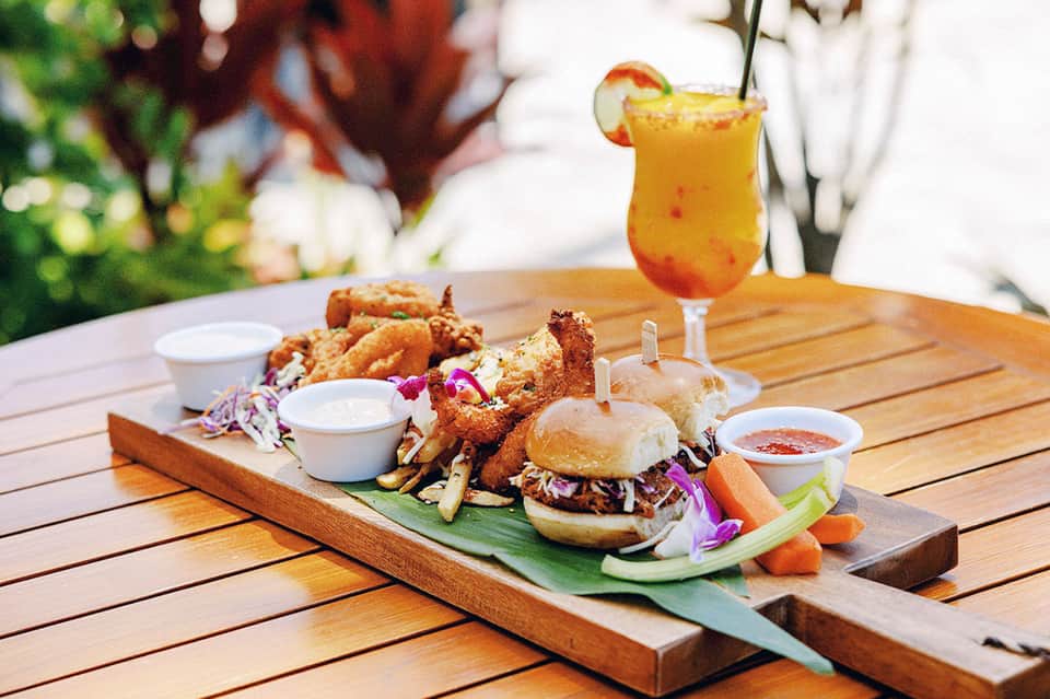 Wooden board of food with burgers, fried chicken, dips, and tropical drink on outdoor table with garden