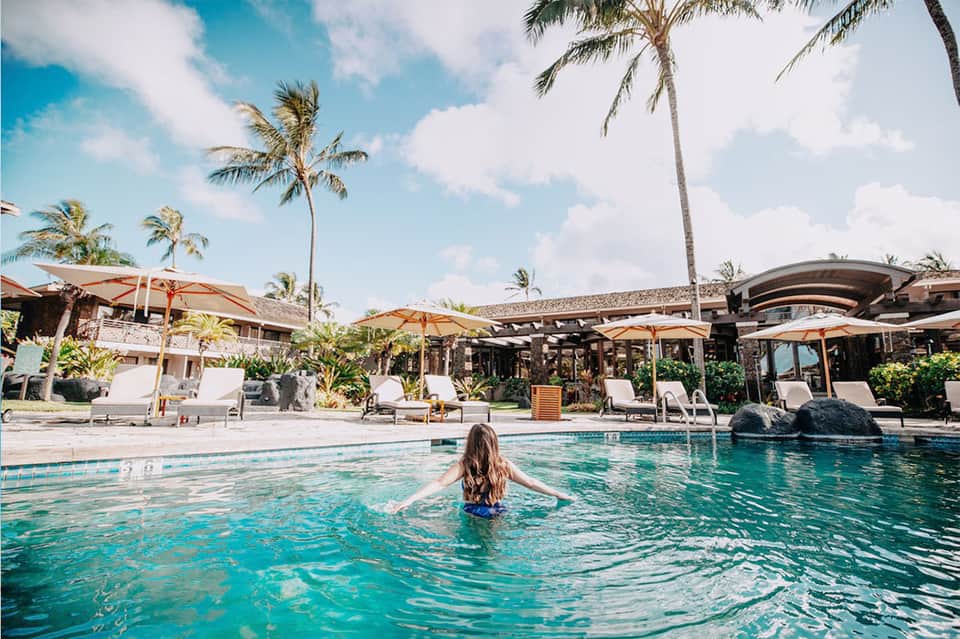Woman swimming in resort pool with palm trees, lounge chairs, and umbrellas in tropical setting
