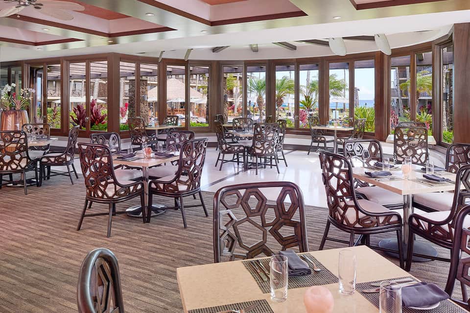 Upscale dining room with ocean view windows, elegant chairs, and tropical flowers overlooking beach