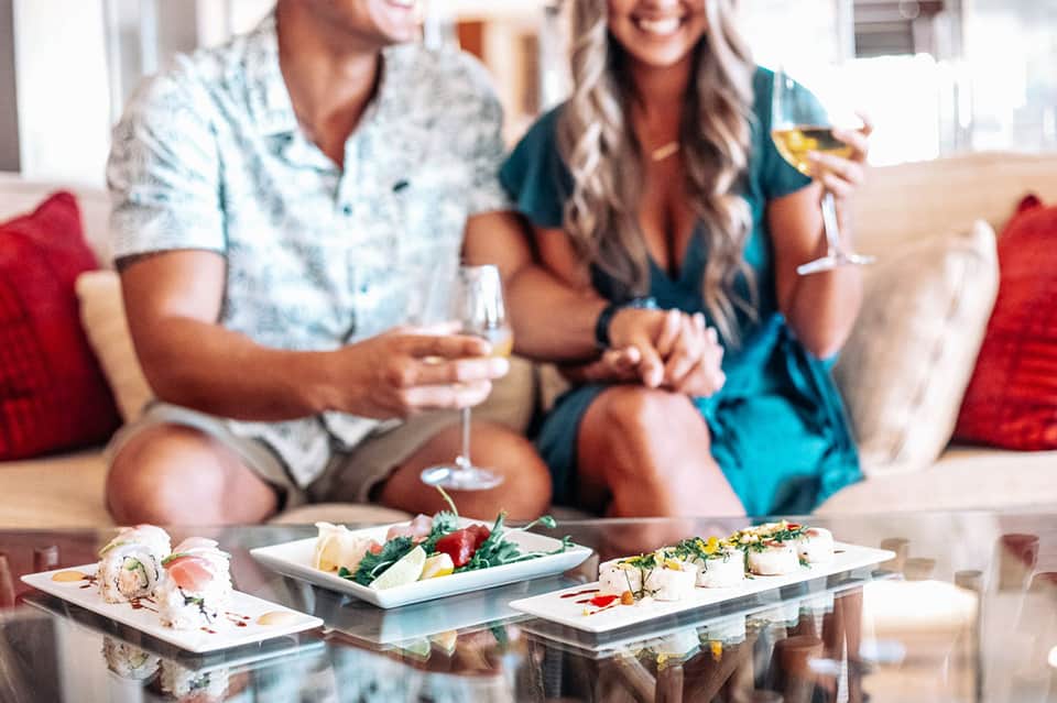 Couple enjoying appetizers and wine on beige sofa with seafood and sushi dishes on glass table