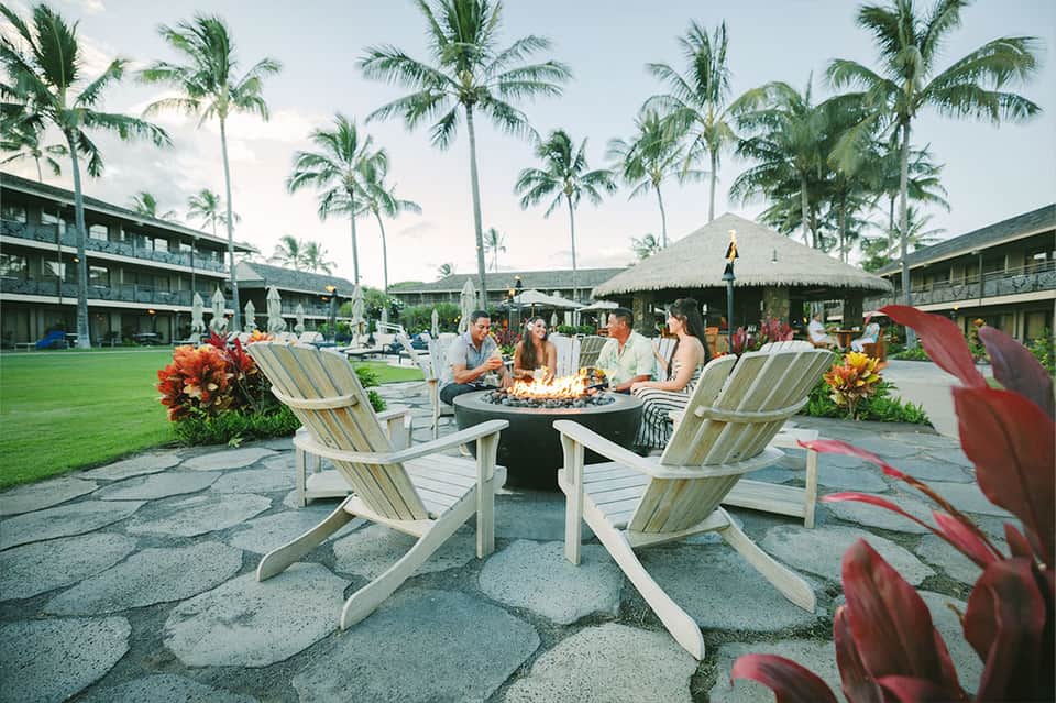 Resort courtyard with fire pit, Adirondack chairs, palm trees, tropical flowers, and resort buildings in background