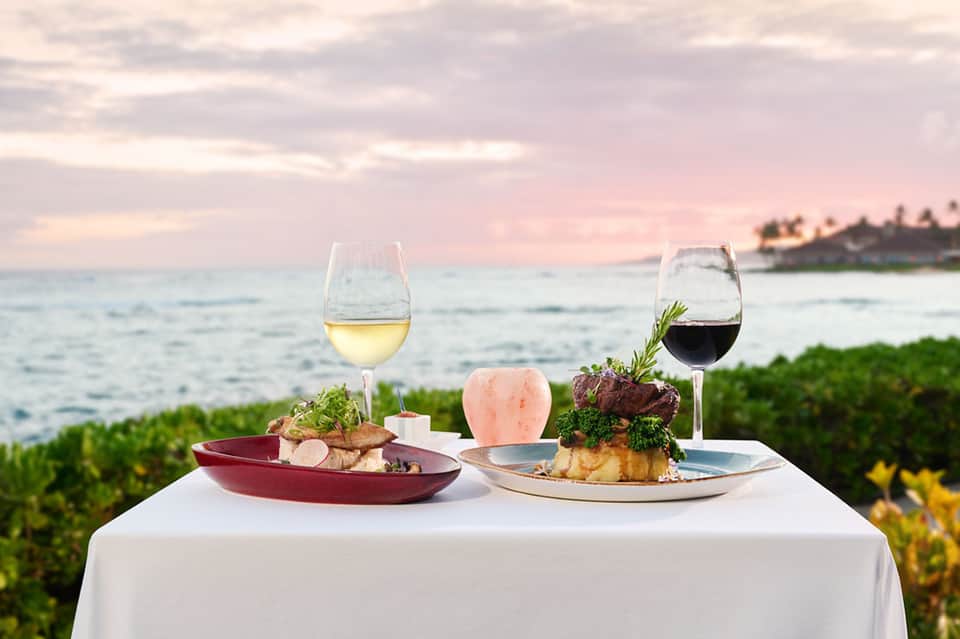 Plated dinner with wine glasses overlooking sunset ocean and tropical foliage