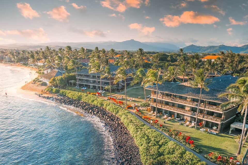 Aerial view of beachfront resort with palm trees, pools, and mountains in distance at sunset