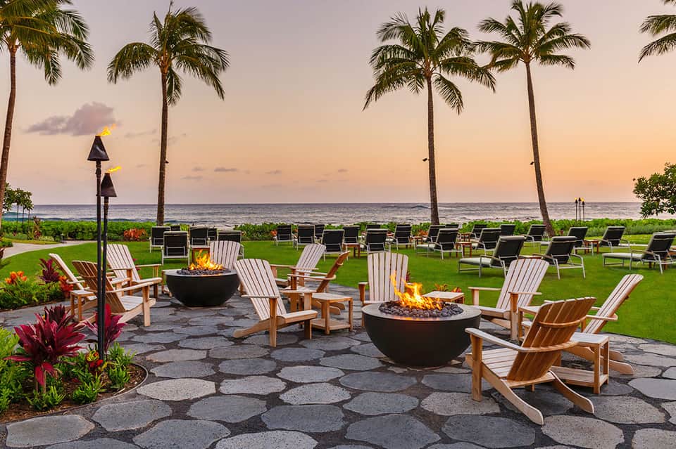 Beachfront fire pit area with Adirondack chairs, palm trees, and ocean sunset view