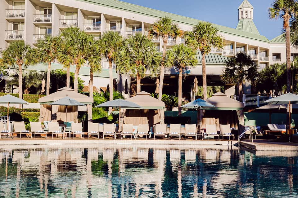Beachfront resort building with palm trees, white umbrellas, and lounge chairs by the pool